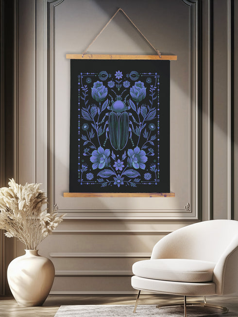 A symmetrical dark wall tapestry featuring a large iridescent blue beetle centered among intricate floral motifs and botanical elements on a black background, suspended from a wooden dowel with twine.